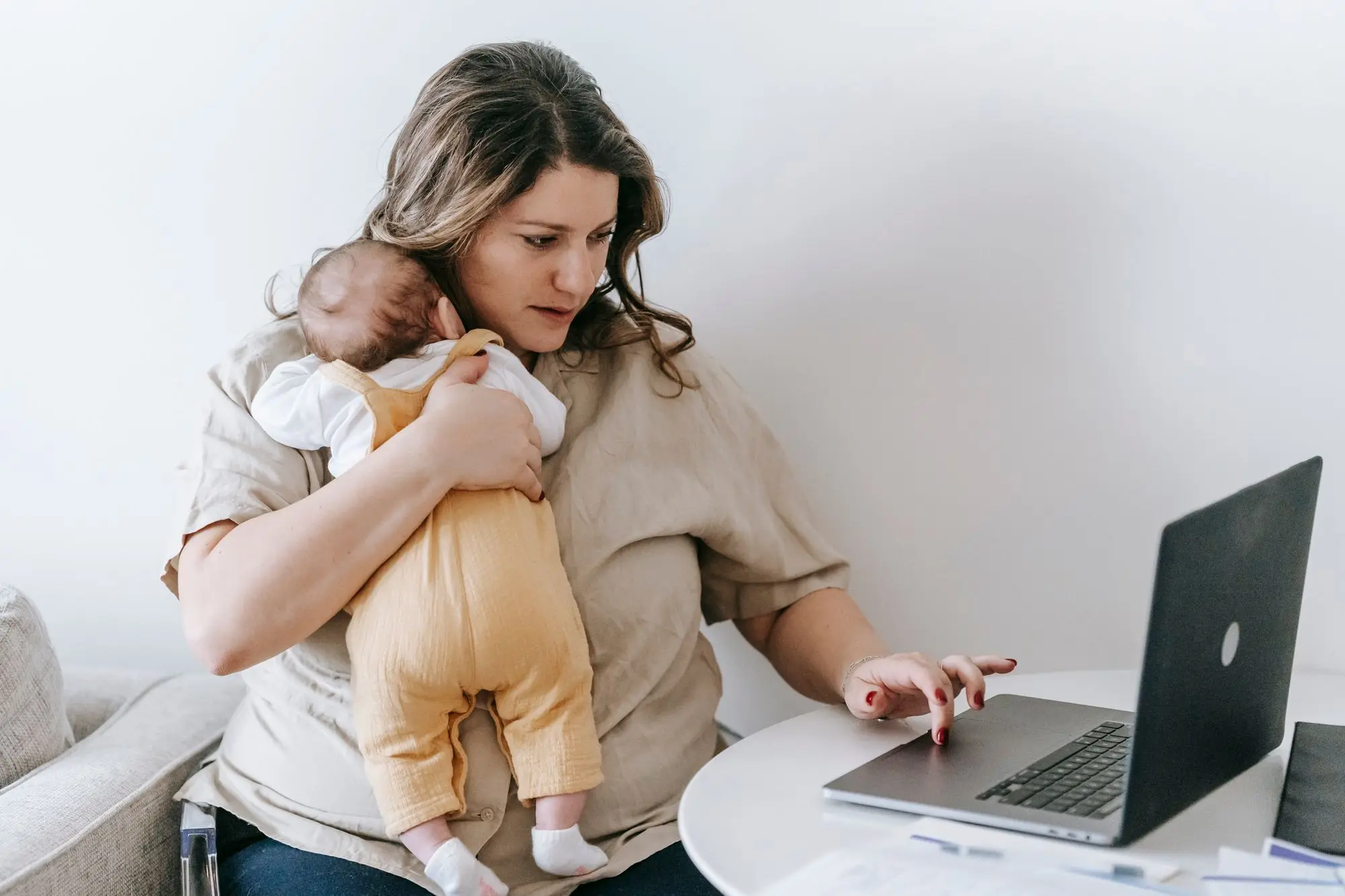 Business owner holding a newborn on her shoulder while checking the business accounts on a laptop at home